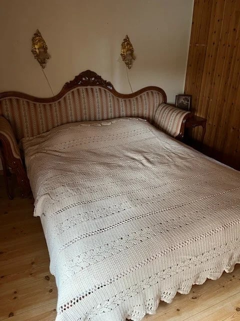 Ornate wooden bed with cream striped upholstered headboard and footboard, topped with white crochet bedspread, in room with wood paneling