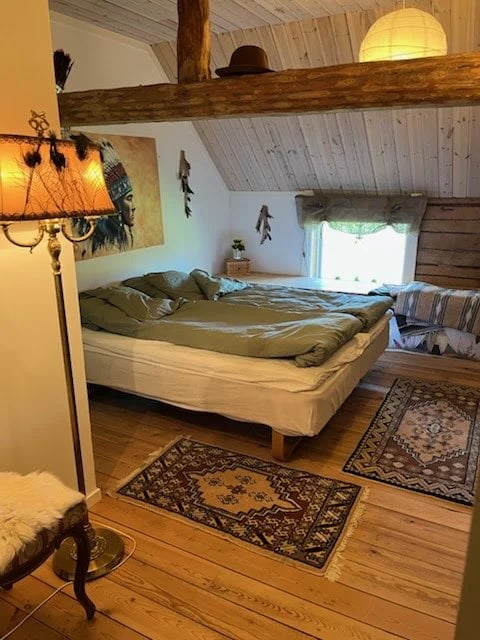 Rustic bedroom loft with exposed wooden beams, queen bed with olive green bedding, decorative floor rugs, and warm lighting