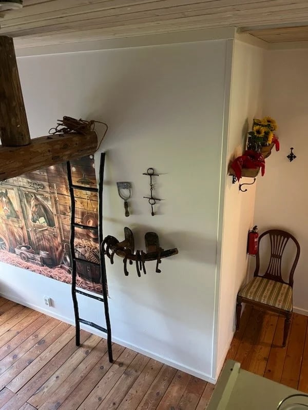 Interior room with white display wall featuring mounted antique tools and metal artifacts, wooden ladder leaning against wall, vintage chair, wooden flooring