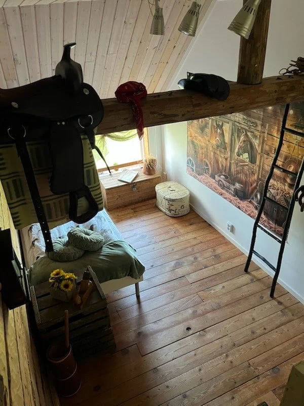 Rustic loft room with wooden beams, saddle and equestrian equipment hung on wall, bed with green bedding, and wooden flooring