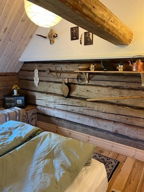 Rustic bedroom with wooden log cabin walls, exposed beam ceiling, bed with green bedding, and shelving with decorative items and hanging accessories
