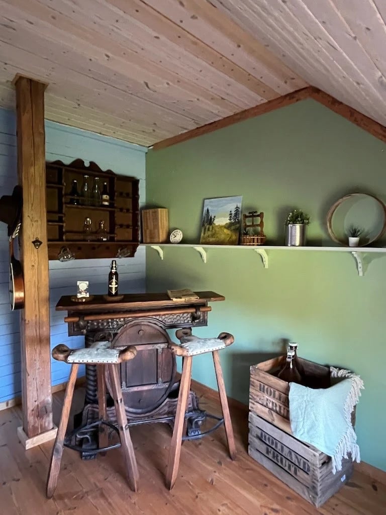 Rustic cabin interior with wooden desk, chair, shelves with decor items on sage green wall, and wooden ceiling beams