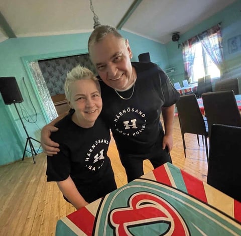 Two people in black matching t-shirts smile while standing together in a turquoise room above a colorful geometric artwork